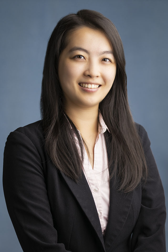 Studio headshot of a smiling woman in a dark blazer against a blue-gray backdrop